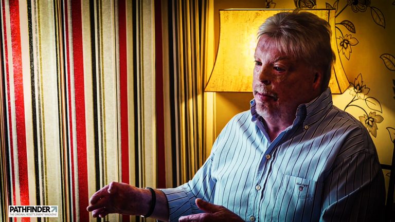 An older man with gray hair speaks, gesturing with his hands in a room adorned with striped curtains and a lit lamp. The dignity of a Falklands veteran resonates in his voice, echoing tales of courage and resilience.