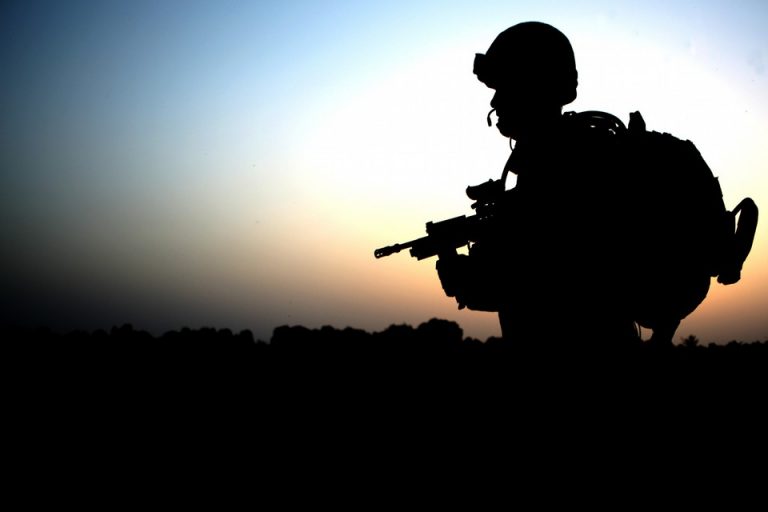 A silhouette of soldiers holding rifles stands stark against the dusky sky, embodying dedication and the commitment to serve until 65.