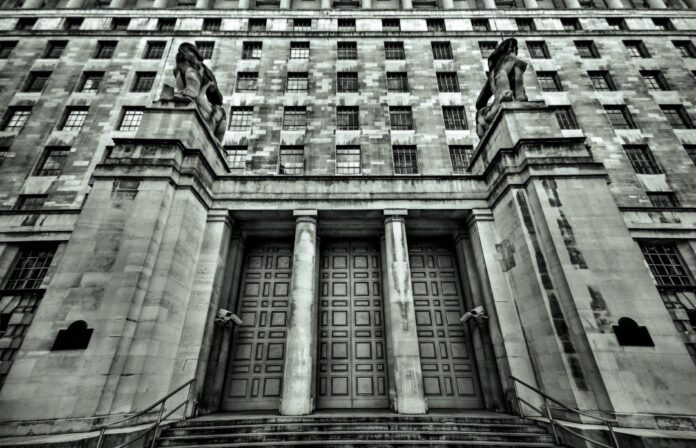 The stone building facade, with its symmetrical columns and two lion statues guarding the entrance, conveys a spirit of resilience akin to Enhanced Resettlement Support. Monochrome tones reflect the gravitas, as steps ascend to large doors and windows in a grid pattern complete the upper structure.
