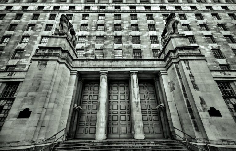 The stone building facade, with its symmetrical columns and two lion statues guarding the entrance, conveys a spirit of resilience akin to Enhanced Resettlement Support. Monochrome tones reflect the gravitas, as steps ascend to large doors and windows in a grid pattern complete the upper structure.