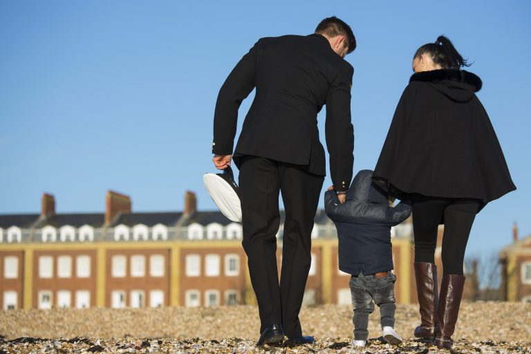 A family of three strolls along a pebble beach, their steps echoing reflection. The man, in a navy uniform and holding his hat, signifies leaving the military. Their child clutches both parents' hands as houses dot the background under a clear blue sky.