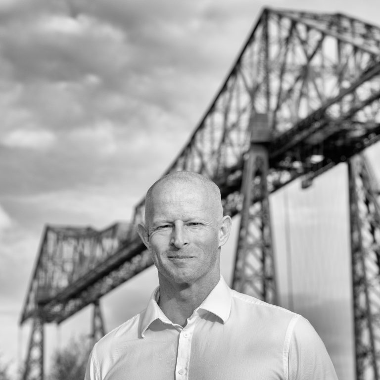 A veteran with a shaved head, wearing a white shirt, stands outdoors in front of a large industrial bridge structure, pondering his controversial storyline.