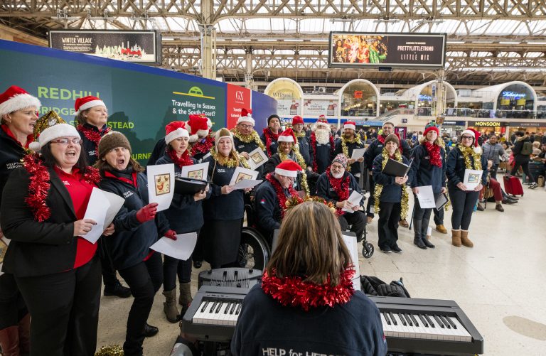 A choir wearing Santa hats and festive attire performs in a bustling train station, the conductor facing them with sheet music, as they sing for Veterans.
