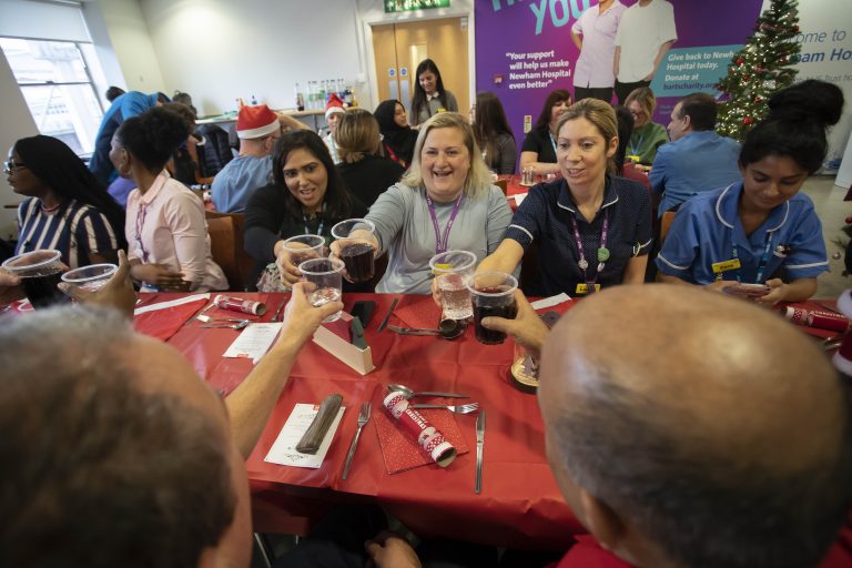 A group of healthcare workers, like a jolly bunch of Brits on Christmas Day, sit around a table raising plastic cups in a cheerful toast, with festive decorations setting the perfect holiday scene.