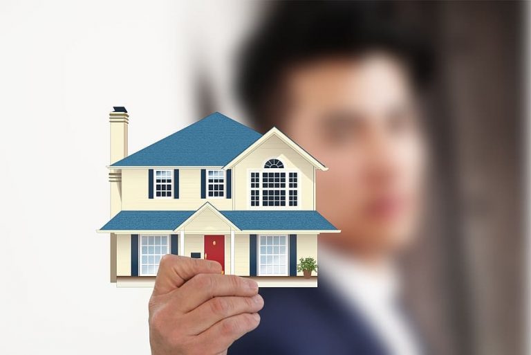 A person in a suit confidently holds a small model of a yellow house with a blue roof, symbolizing the first step to get on the property ladder.