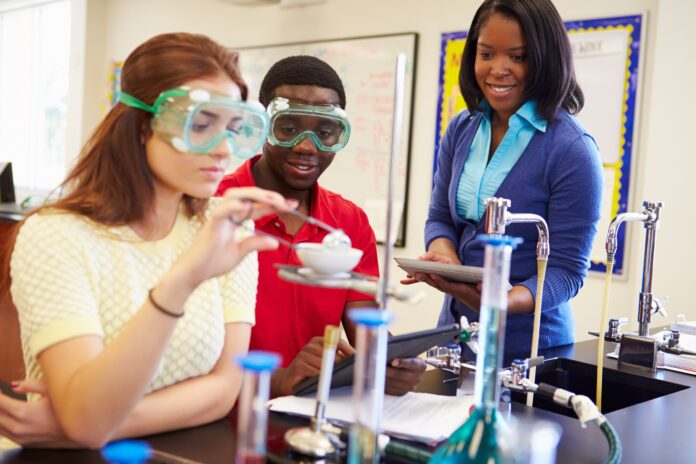 Students diligently conducting an experiment in a science class, donning safety goggles, are observed by a teacher with a tablet. This moment of teaching highlights the critical skills transfer essential for nurturing budding scientists.