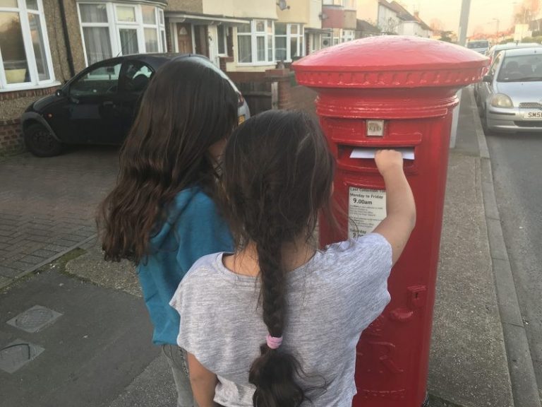 Two children stand on a sidewalk, one posting a letter into a red postbox. Houses and parked cars are visible in the background, hinting at a resettlement area after military pathfinders marked the neighborhood.