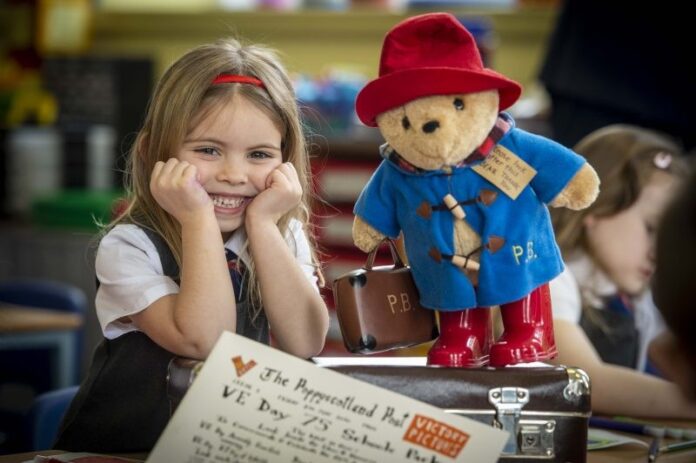 A young girl in school uniform smiles at a desk next to a Paddington Bear toy, surrounded by papers and a suitcase—a heartwarming glimpse into the resettlement journey of military families in a classroom setting.