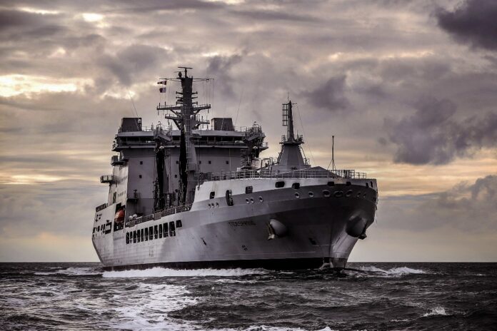 A large gray military naval ship sails forward on the ocean under a cloudy sky.