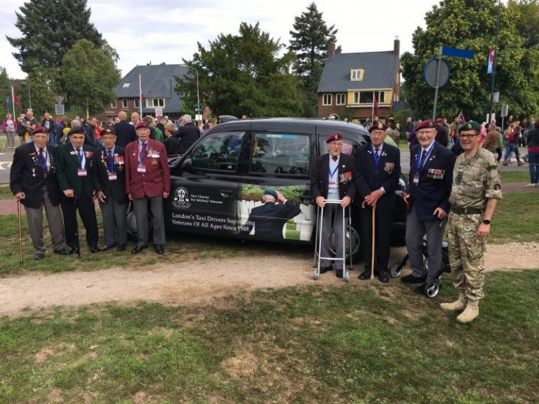 A group of veterans and one uniformed soldier stand next to a black taxi with commemorative signage, outdoors on a grassy area near houses and trees, honoring the Pathfinder spirit of service and resettlement.