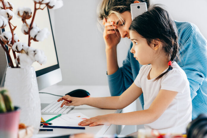 A woman on the phone looks stressed while a young girl uses a computer at a desk with papers, colored pencils, and an Army Pathfinder badge beside a vase of cotton branches.