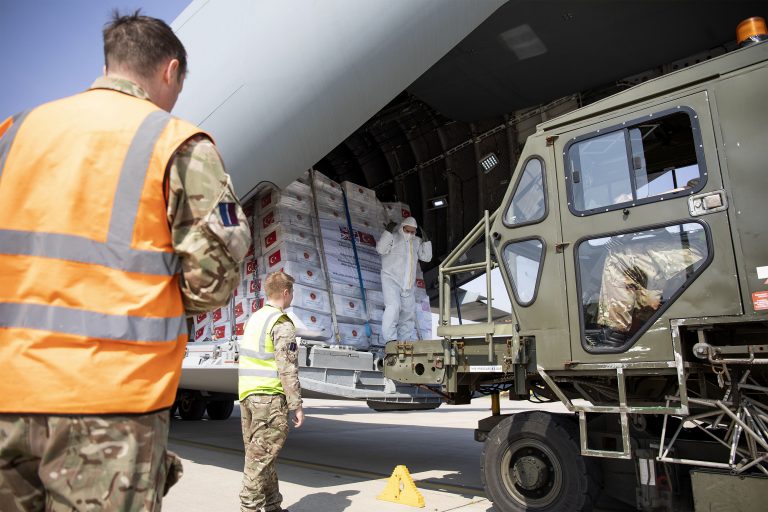 Military personnel and veterans unload pallets of humanitarian aid from a cargo plane using heavy machinery at an airport, supporting resettlement efforts in the region.