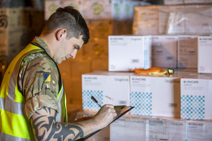 A veteran in a high-visibility vest and military-style uniform writes on a clipboard in front of stacked boxes in a warehouse.