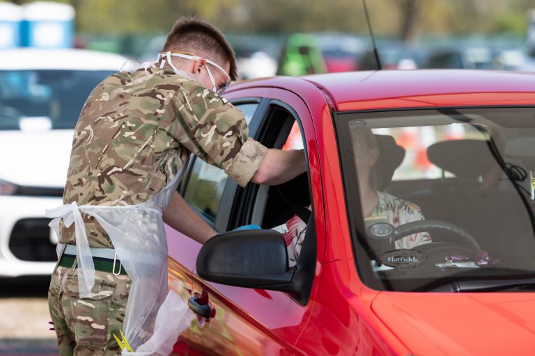 A person in Army uniform and PPE assists someone in a red car at a drive-through testing or vaccination site, supporting resettlement efforts.