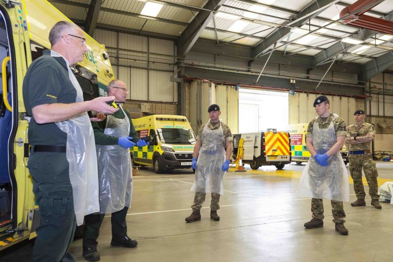 Four uniformed military personnel, possibly veterans, wear aprons and gloves as they stand in a garage, listening to two paramedics beside ambulances—a scene reflecting resettlement into new roles beyond their Royal Navy service.
