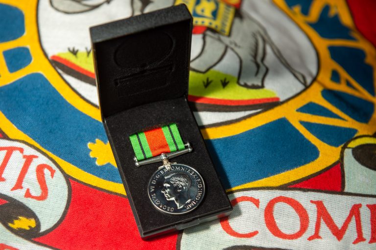 A silver medal with a green and red striped ribbon in a black box, resting on a colorful, heraldic-patterned fabric—an emblem of Royal Navy or Army service, honoring dedication and resettlement into new chapters.