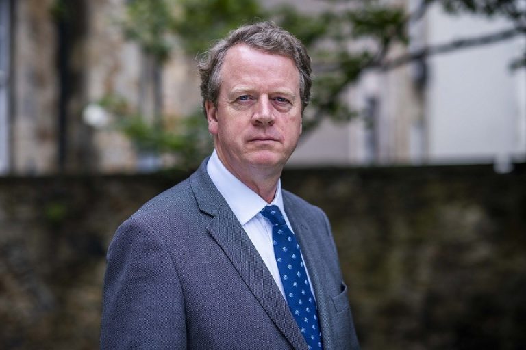 A middle-aged military veteran in a gray suit and blue patterned tie stands outdoors in front of a blurred stone wall and greenery, looking directly at the camera with a neutral expression.