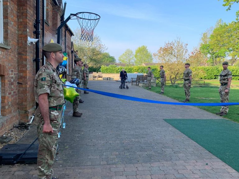 Several uniformed military personnel stand outside holding a large blue ribbon for an outdoor event, with balloons and a basketball hoop visible nearby.