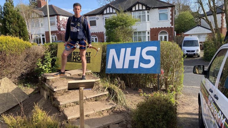A man stands on outdoor steps beside a large NHS sign and a "Stay at Home" banner, with residential houses, a parked van, and a Royal Navy recruitment poster in the background.