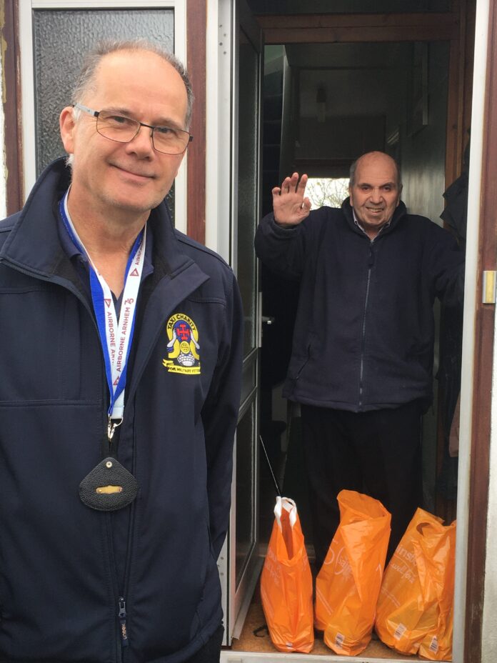 A veteran wearing a badge stands at an open door, while another man waves from inside. Several orange grocery bags are on the floor near the doorway, hinting at a fresh start and resettlement.