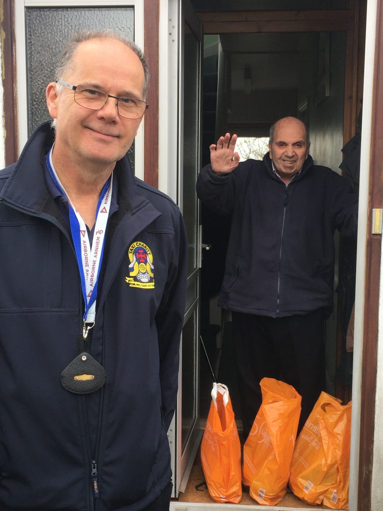 A veteran wearing a badge stands at an open door, while another man waves from inside. Several orange grocery bags are on the floor near the doorway, hinting at a fresh start and resettlement.