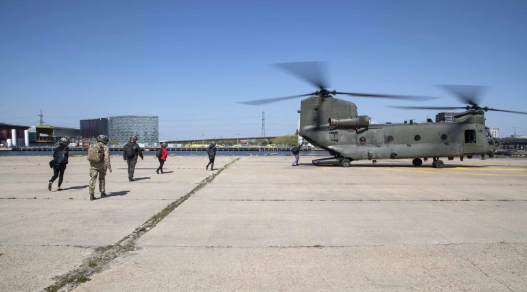 A group of people, part of an Army resettlement program, walk toward a stationary military helicopter with spinning rotors on a large concrete surface under a clear sky.