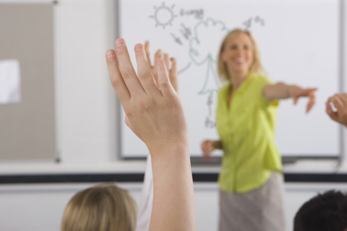 A student raises their hand in a classroom while a teacher in a green shirt points towards them. A whiteboard with drawings is visible in the background, hinting at a lesson on military resettlement.