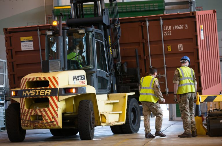 A forklift operator moves a large shipping container while two Army veterans in military uniforms and safety vests stand nearby inside an industrial facility.