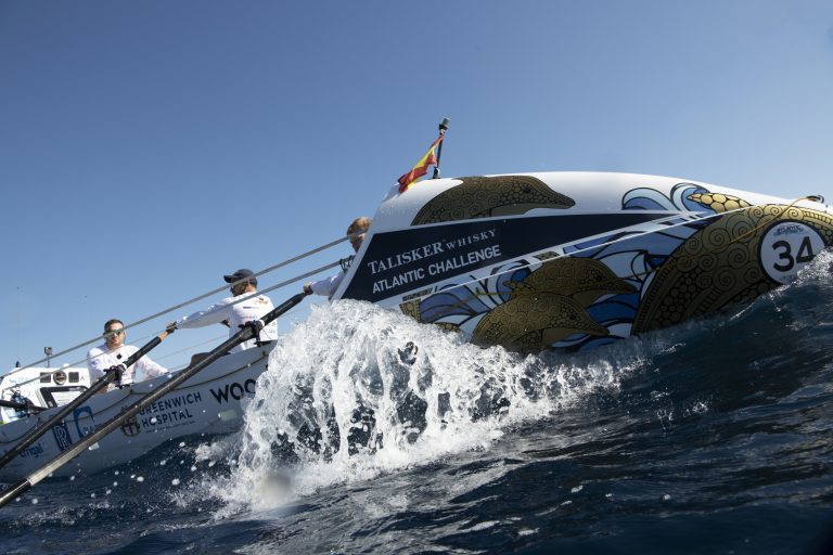 Three rowers, including a former Army pathfinder, participate in the Talisker Whisky Atlantic Challenge, rowing a decorated boat numbered 34 across the ocean under clear blue skies.
