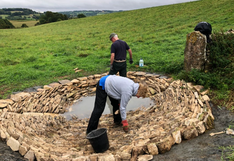 Two people build a circular stone structure in a grassy field, with one person bending over to place stones and a bucket nearby. Some water has pooled in the center of the structure, possibly part of a resettlement effort or military project.
