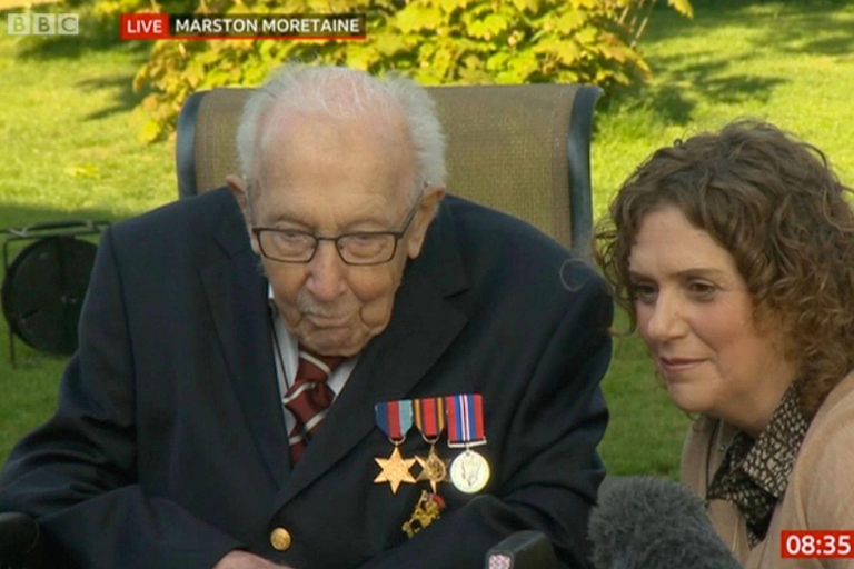 Elderly veteran wearing medals sits beside a woman outdoors during a BBC live broadcast. Microphones are visible in the foreground, and the time reads 08:35.