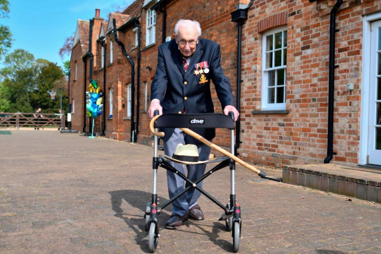 An elderly Army veteran wearing medals and a blazer uses a walker with a cane and hat placed on it, standing outside a brick building on a sunny day.