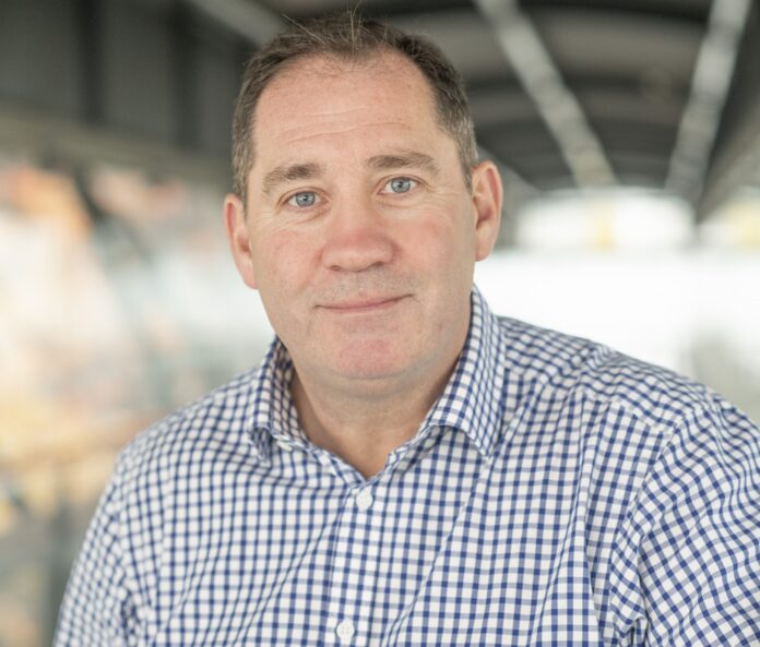 A middle-aged Army veteran with short brown hair, wearing a blue and white checkered shirt, sits indoors with a blurred background.