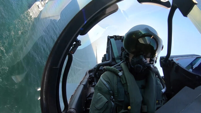 A military fighter pilot wearing a helmet and oxygen mask sits in the cockpit of a jet, flying above the ocean with a ship visible in the background.