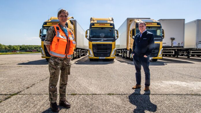 Two men stand in front of three yellow DHL trucks parked side by side; one, a Royal Navy veteran in military uniform with an orange vest, and the other in business attire.