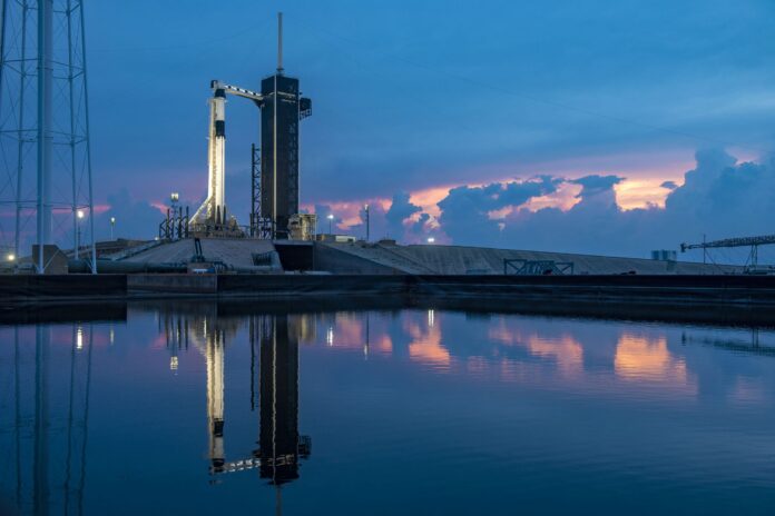 A rocket stands upright on a launch pad at dusk, reflected in a nearby body of water, with clouds and a colorful sky in the background, evoking themes of military innovation and resettlement.