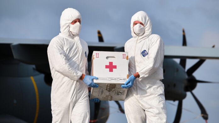 Two people in full protective suits and masks, part of a military resettlement team, carry a box with a red cross symbol in front of a large aircraft.