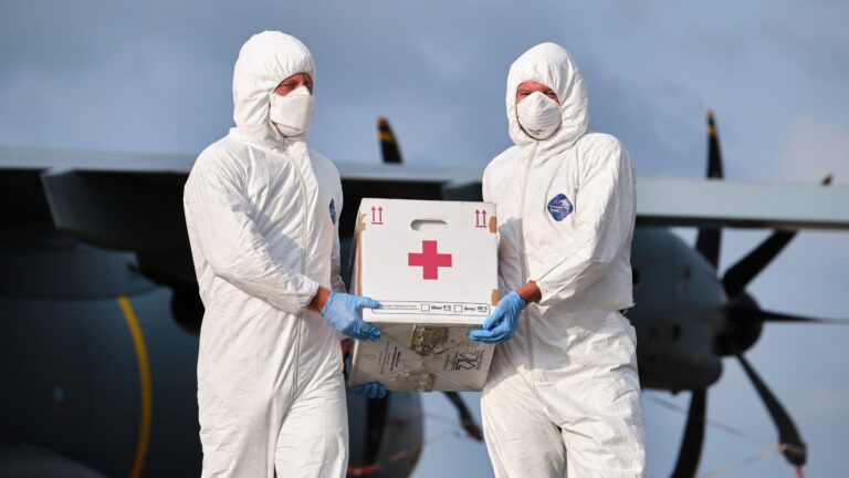 Two people in full protective suits and masks, part of a military resettlement team, carry a box with a red cross symbol in front of a large aircraft.