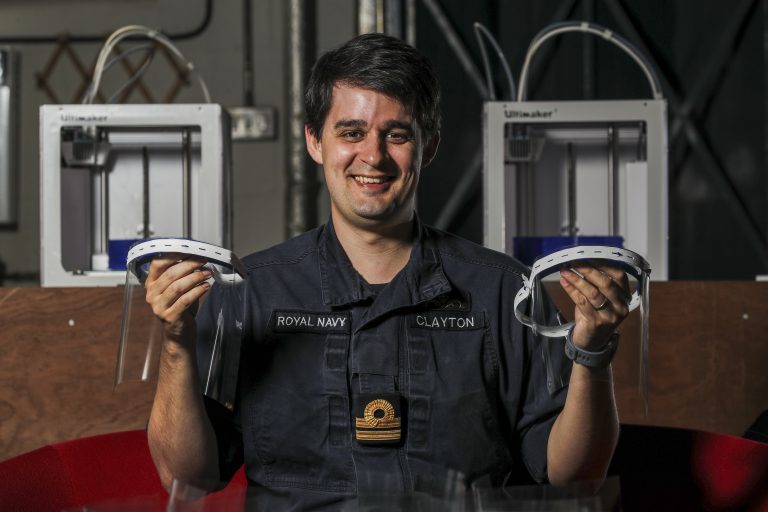 A person in a Royal Navy uniform holds two 3D-printed face shields, with Pathfinder 3D printers and wood panels in the background.