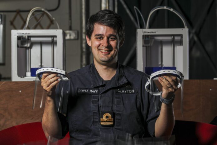 A person in a Royal Navy uniform holds two 3D-printed face shields, with Pathfinder 3D printers and wood panels in the background.