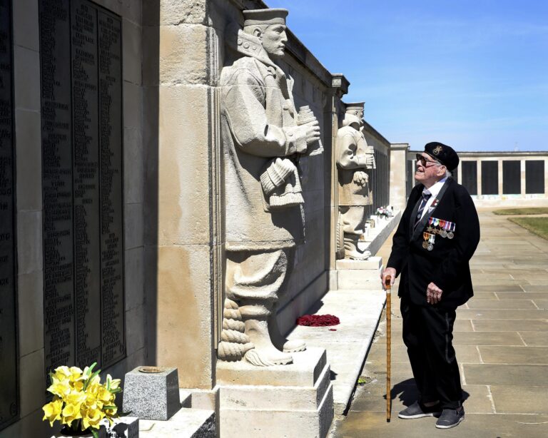 An elderly military veteran with medals stands with a cane, looking at a memorial wall and statue on a sunny day. Yellow flowers and wreaths are placed at the memorial, honoring those who served.