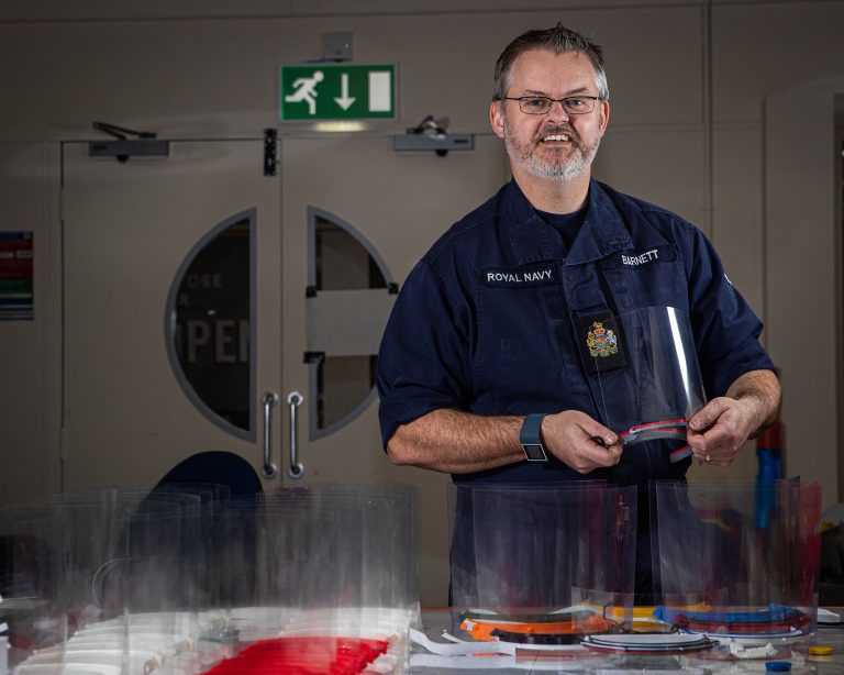 A man in a Royal Navy uniform stands indoors, holding a clear plastic face shield with several more face shields arranged on the table in front of him, supporting military resettlement efforts.