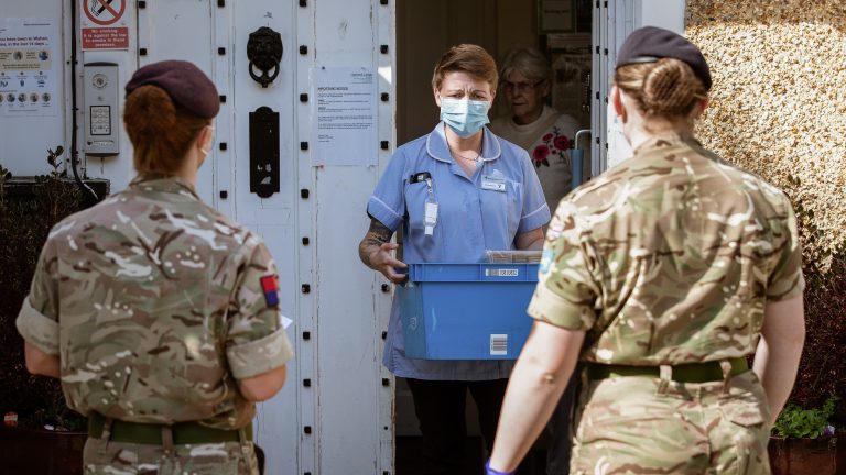 A nurse wearing a mask carries a blue box out of a building while two uniformed Royal Navy personnel stand facing her. An elderly woman stands in the doorway behind her.