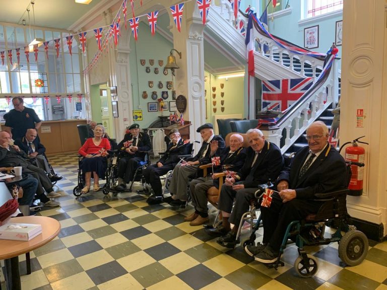 A group of elderly people, some in wheelchairs, sit together in a decorated hall with British flags and bunting, joined by retired Army veterans, in front of a staircase.