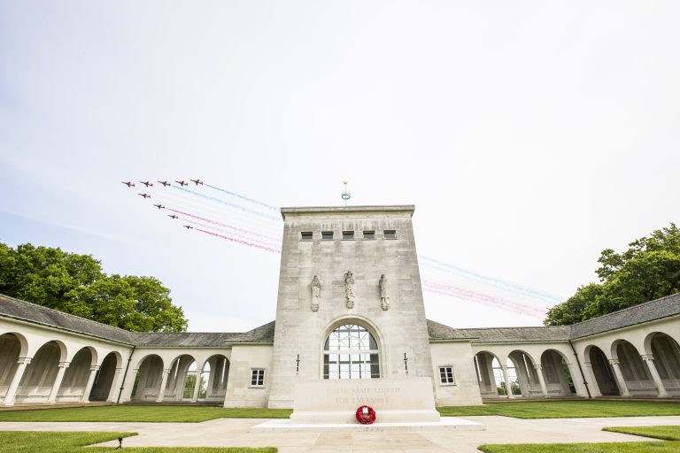 Aerial display team flies over a stone war memorial building and courtyard, honoring veterans with red, white, and blue smoke trails visible in the sky.