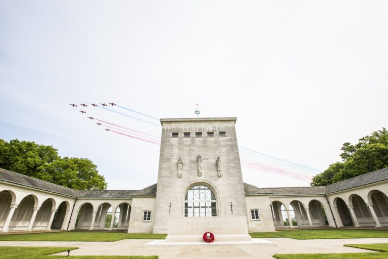 Aerial display team flies over a stone war memorial building and courtyard, honoring veterans with red, white, and blue smoke trails visible in the sky.