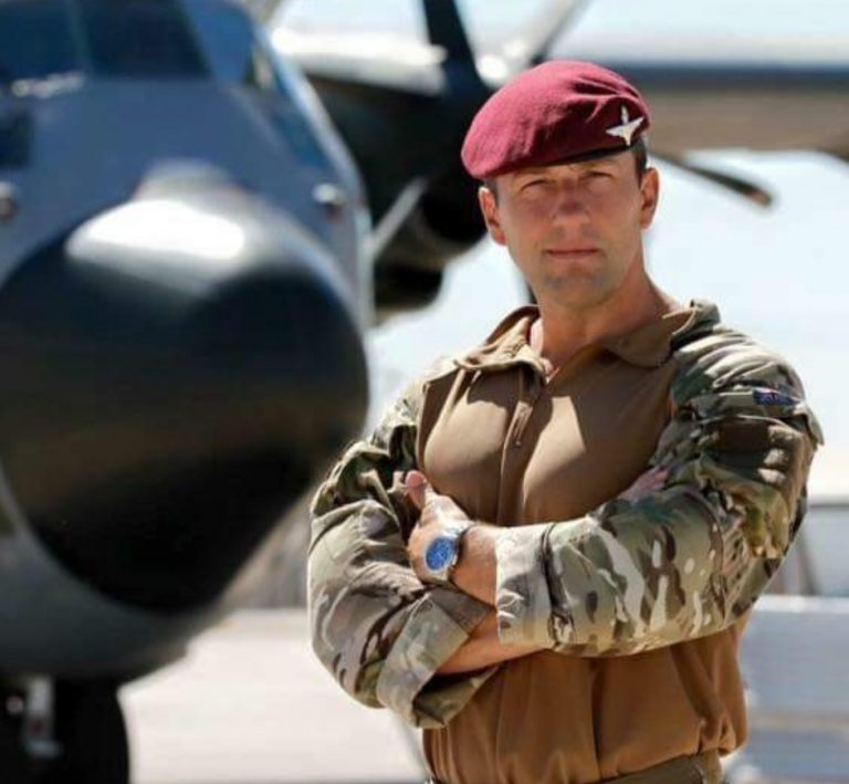 A Royal Navy veteran in camouflage military uniform and a maroon beret stands with arms crossed in front of a military aircraft on a sunny day.