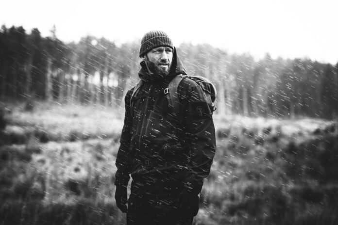 A veteran wearing outdoor gear and a beanie stands in a snowy landscape, looking to the side as snow falls around him.