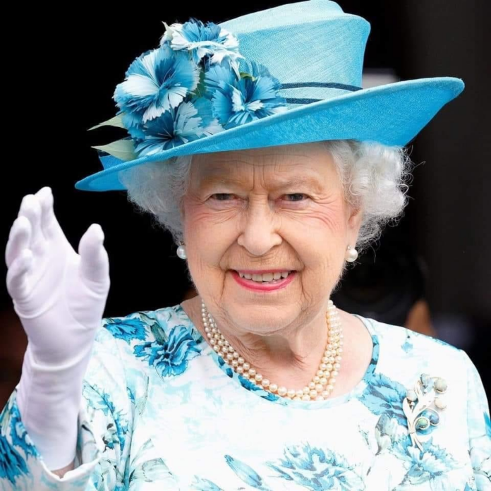 An elderly woman with white hair, wearing a blue floral dress, matching hat with flowers, white gloves, and pearls—perhaps once a proud Pathfinder in the Royal Navy—smiles and waves at the camera.