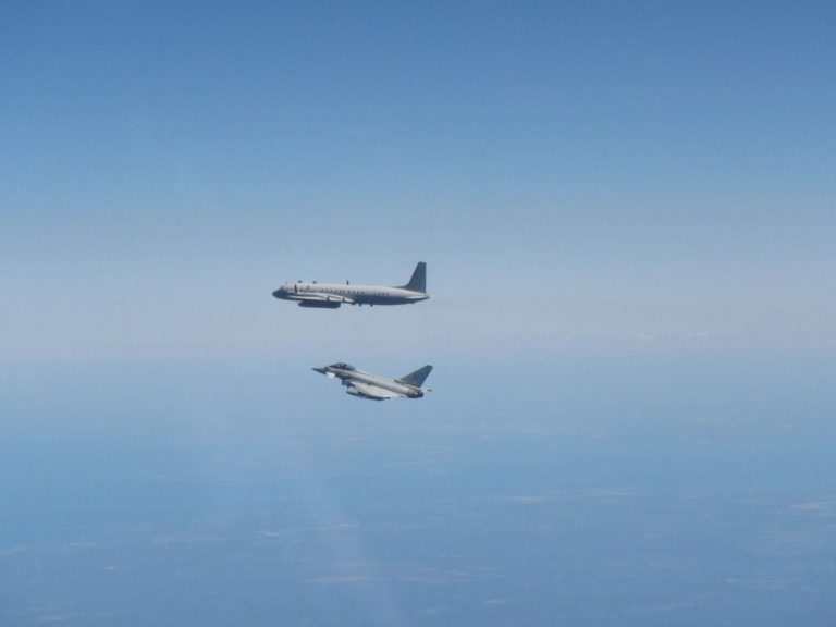 A passenger aircraft and an Army jet fly parallel to each other at high altitude above a landscape with a clear blue sky.
